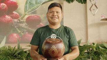 Homem de camiseta verde segura recipiente arredondado de cerâmica com desenhos geométricos, diante de painel com frutos de café.