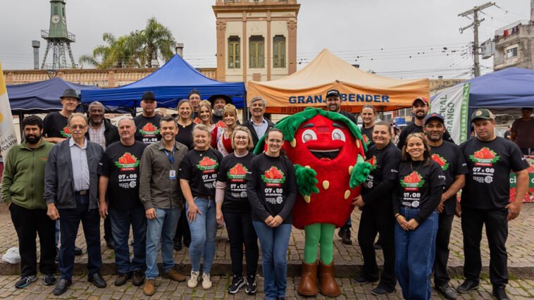 Grupo de organizadores posa ao lado de mascote de morango em frente a barracas de feira na cidade de Pelotas.