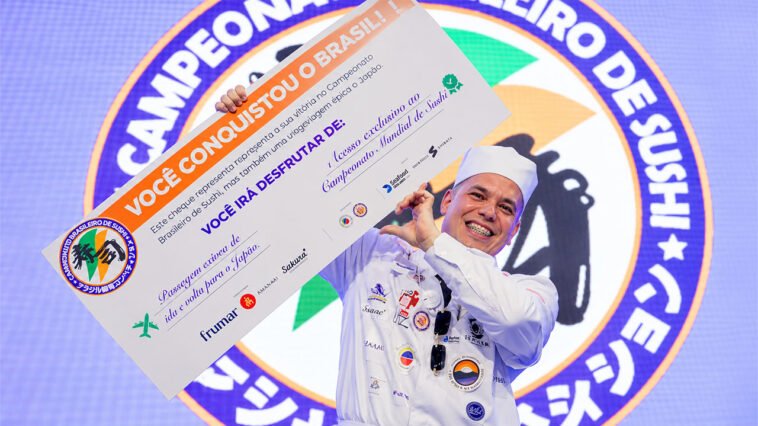 Chef de uniforme branco sorri enquanto ergue prêmio de viagem ao Japão diante de painel do Campeonato Brasileiro de Sushi.