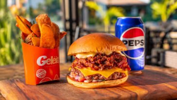 Hambúrguer com carne desfiada e queijo, acompanhado de batatas rústicas em embalagem Geléia e lata de Pepsi.
