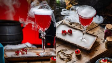 Duas taças de coquetel vermelho envoltas em fumaça branca, com doces em formato de olhos sobre mesa de madeira.