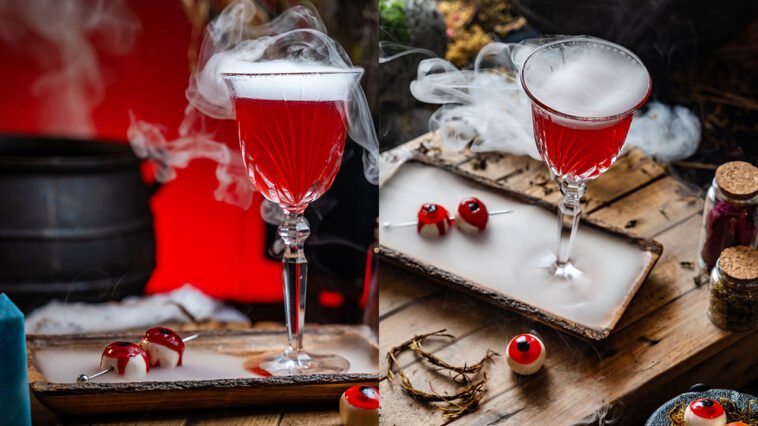 Duas taças de coquetel vermelho envoltas em fumaça branca, com doces em formato de olhos sobre mesa de madeira.
