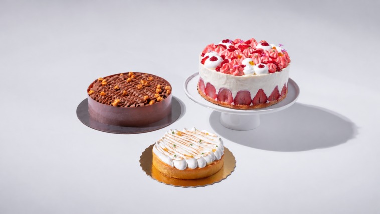Três bolos sobre fundo claro: um de chocolate, outro coberto com creme branco e morangos e um menor com cobertura branca.