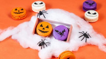 Macarons laranja, roxos e brancos decorados com rostos de abóbora, morcegos e caveiras, entre teias falsas e aranhas pretas.