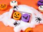 Macarons laranja, roxos e brancos decorados com rostos de ab&oacute;bora, morcegos e caveiras, entre teias falsas e aranhas pretas.