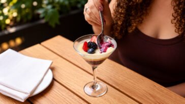 Pessoa leva uma colher à taça com sobremesa de creme claro, frutas vermelhas e base de chocolate sobre mesa de madeira.