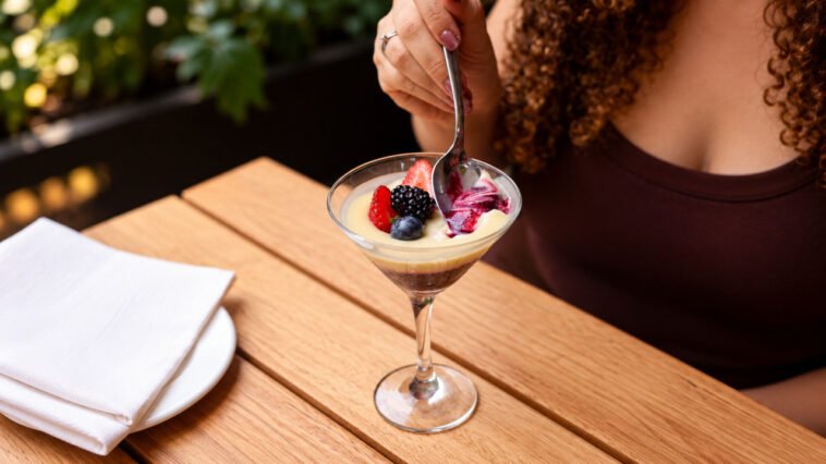 Pessoa leva uma colher à taça com sobremesa de creme claro, frutas vermelhas e base de chocolate sobre mesa de madeira.