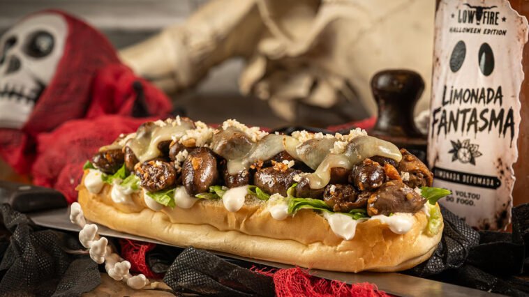 Sanduíche comprido com carne confitada, folhas, molho claro e queijo derretido, ao lado de decoração de Halloween e placa Limonada Fantasma.