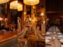 Montagem de ambientes do Maison Dodoma com bar iluminado, figuras decorativas espelhadas e mesa comprida posta para jantar.