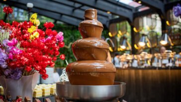 Fonte de chocolate de três níveis ao lado de flores coloridas e mesa de sobremesas em ambiente de brunch.