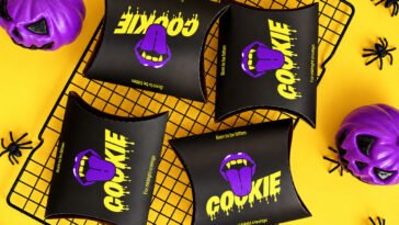 Embalagens pretas com a palavra Cookie em amarelo e boca roxa, cercadas por abóboras roxas e aranhas decorativas.