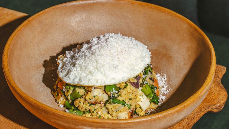 Prato fundo de cerâmica com legumes, farofa e ervas, coberto por preparação branca oval com raspas claras.