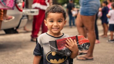 Menino sorridente segura caminhão de brinquedo vermelho em ambiente com outras pessoas ao fundo.