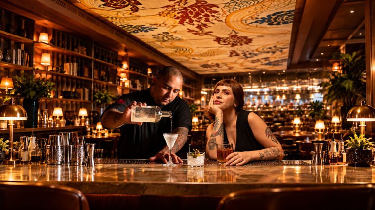 Bartender serve bebida em taça no balcão enquanto mulher observa, em bar com prateleiras iluminadas.
