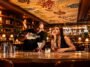 Bartender serve bebida em ta&ccedil;a no balc&atilde;o enquanto mulher observa, em bar com prateleiras iluminadas.