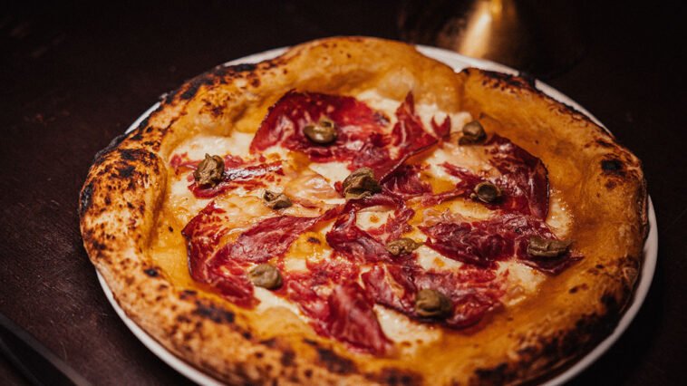 Pizza de borda alta e tostada, coberta com queijo derretido, fatias finas de carne curada e pequenos pedaços de cogumelos.