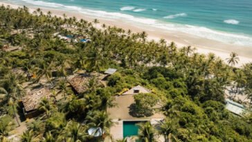 Vista a&eacute;rea de bangal&ocirc;s e piscina entre coqueiros, ao lado de praia de areia clara e mar azul.